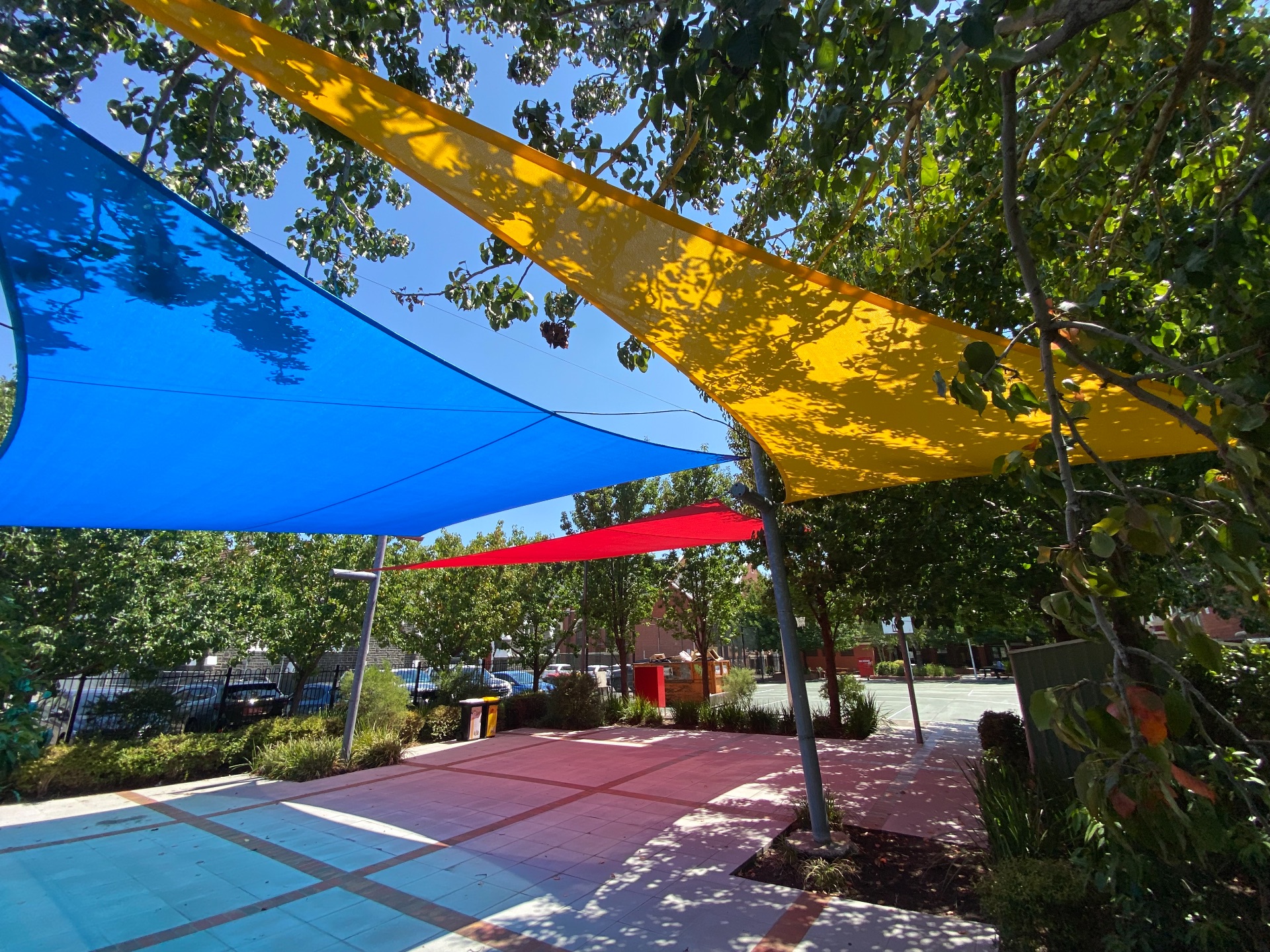 School Shade Sails over a playground sports court. The sails are blue, yellow and red and casting shadows over the court.